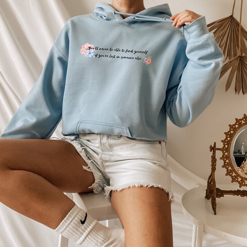 Peut inclure: Un sweat &agrave; capuche bleu clair avec le texte "You'll never be able to find yourself if you're lost in someone else" et un motif floral aquarelle.