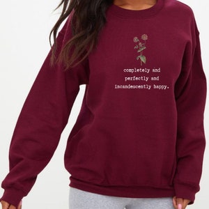 Puede incluir: Sudadera color burdeos con un peque&ntilde;o gr&aacute;fico floral y el texto "completely and perfectly and incandescently happy."