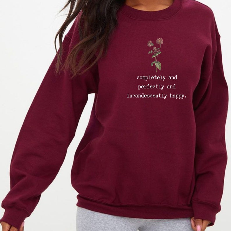 Puede incluir: Sudadera color burdeos con un peque&ntilde;o gr&aacute;fico floral y el texto "completely and perfectly and incandescently happy."