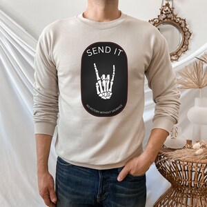 May include: A beige sweatshirt with a black oval graphic featuring a white skeleton hand making a rock and roll sign. The text "SEND IT" is above the hand and "NO VICTORY WITHOUT SACRIFICE" is below the hand.