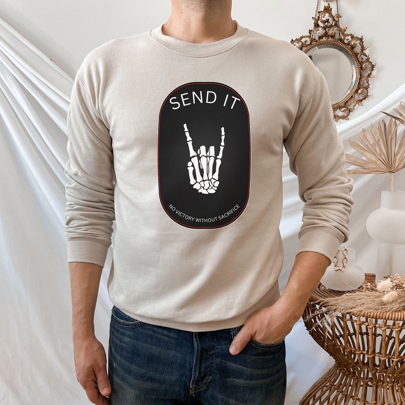May include: A beige sweatshirt with a black oval graphic featuring a white skeleton hand making a rock and roll sign. The text "SEND IT" is above the hand and "NO VICTORY WITHOUT SACRIFICE" is below the hand.