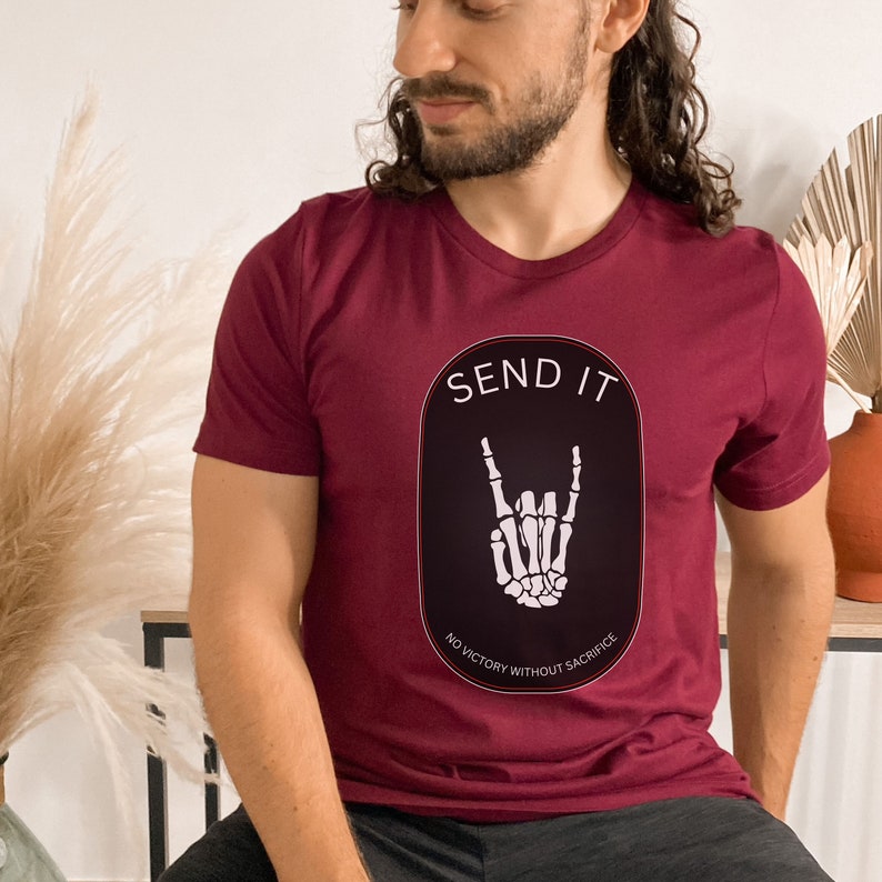 May include: A maroon t-shirt with a black oval graphic featuring a skeleton hand making the rock and roll sign. The text "SEND IT" is above the hand and "NO VICTORY WITHOUT SACRIFICE" is below the hand.