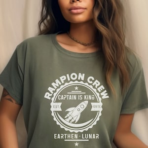 May include: A green t-shirt with a white graphic of a rocket ship and the text "Rampion Crew, Captain is King, Est. 126 T.E., Earthen - Lunar".