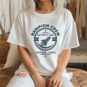 May include: White t-shirt with a navy blue graphic of a rocket ship and the text "Rampion Crew, Captain is King, Est. 128TE, Earthen - Lunar".