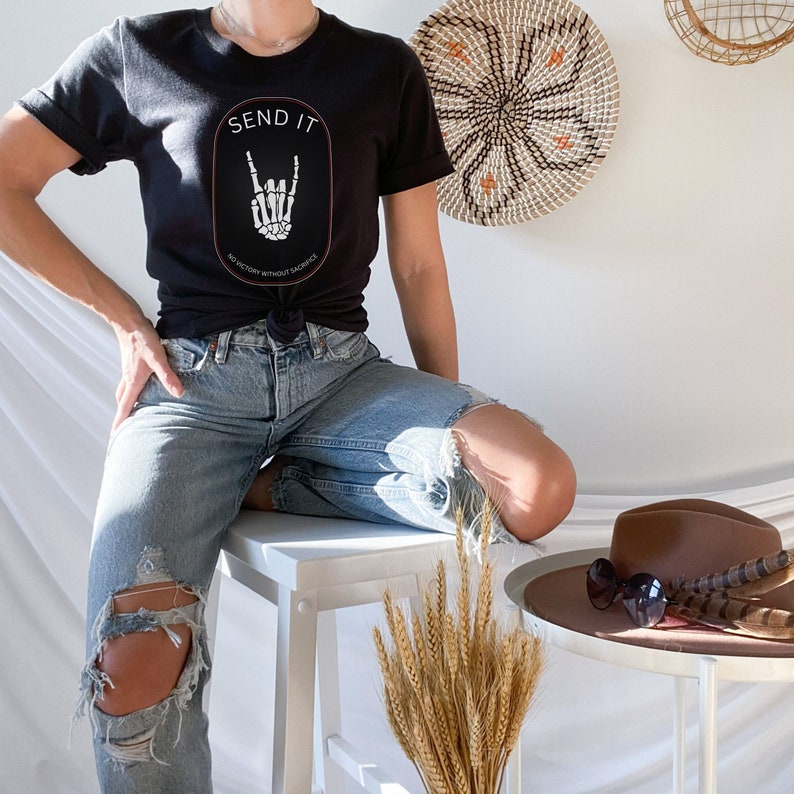 May include: A black t-shirt with a white graphic of a skeleton hand making a rock and roll sign. The graphic is inside a circle with the text "SEND IT" above the hand and "NO VICTORY WITHOUT SACRIFICE" below the hand. The person wearing the shirt is sitting on a white stool with ripped jeans.