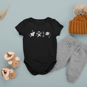 May include: Black baby onesie with a white astronaut, rocket, and planets design. The onesie is paired with gray sweatpants, a brown knit beanie with a white pom pom, and brown baby booties.