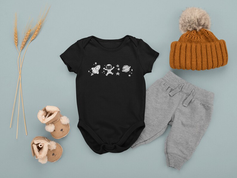 May include: Black baby onesie with a white astronaut, rocket, and planets design. The onesie is paired with gray sweatpants, a brown knit beanie with a white pom pom, and brown baby booties.
