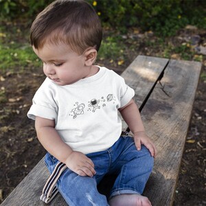 May include: A young child wearing a white t-shirt with a black and white illustration of an astronaut, a rocket ship, and planets. The child is sitting on a wooden bench.