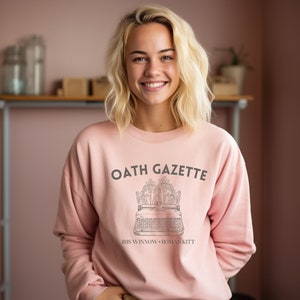 Puede incluir: Una sudadera rosa con el texto "OATH GAZETTE" impreso en la parte delantera. El texto est&aacute; en fuente negra y est&aacute; rodeado por un dise&ntilde;o floral. Debajo del texto est&aacute; el texto "IRIS WINNOW &bull; ROMAN KITT" en una fuente m&aacute;s peque&ntilde;a.