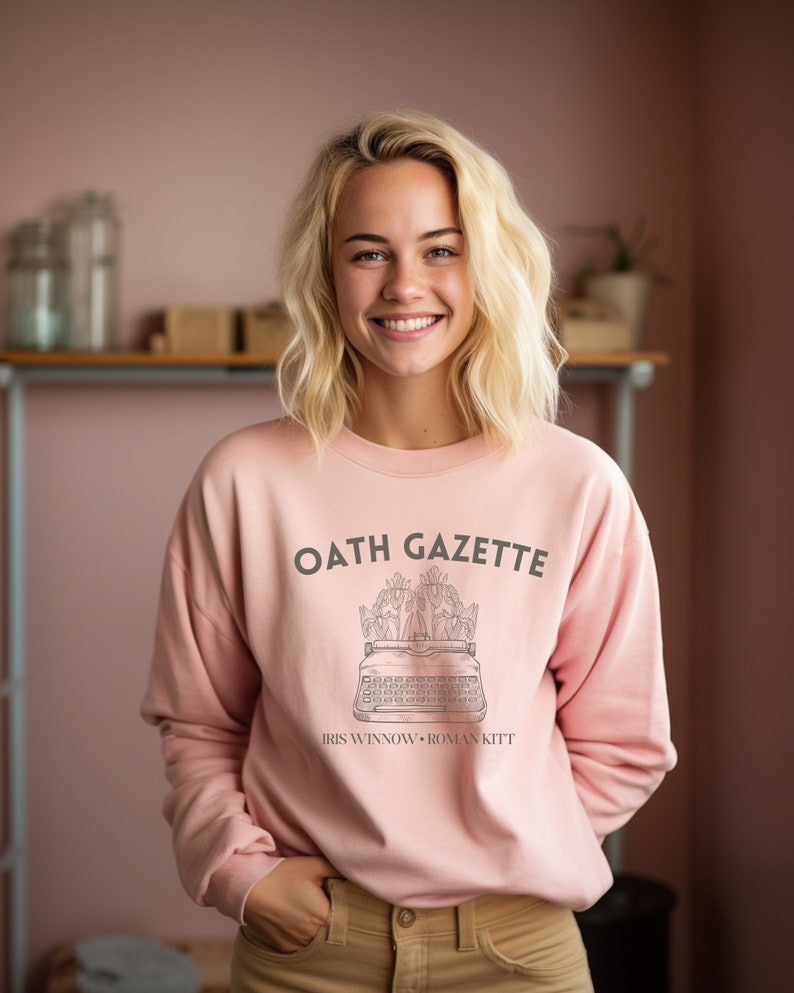 Puede incluir: Una sudadera rosa con el texto "OATH GAZETTE" impreso en la parte delantera. El texto est&aacute; en fuente negra y est&aacute; rodeado por un dise&ntilde;o floral. Debajo del texto est&aacute; el texto "IRIS WINNOW &bull; ROMAN KITT" en una fuente m&aacute;s peque&ntilde;a.