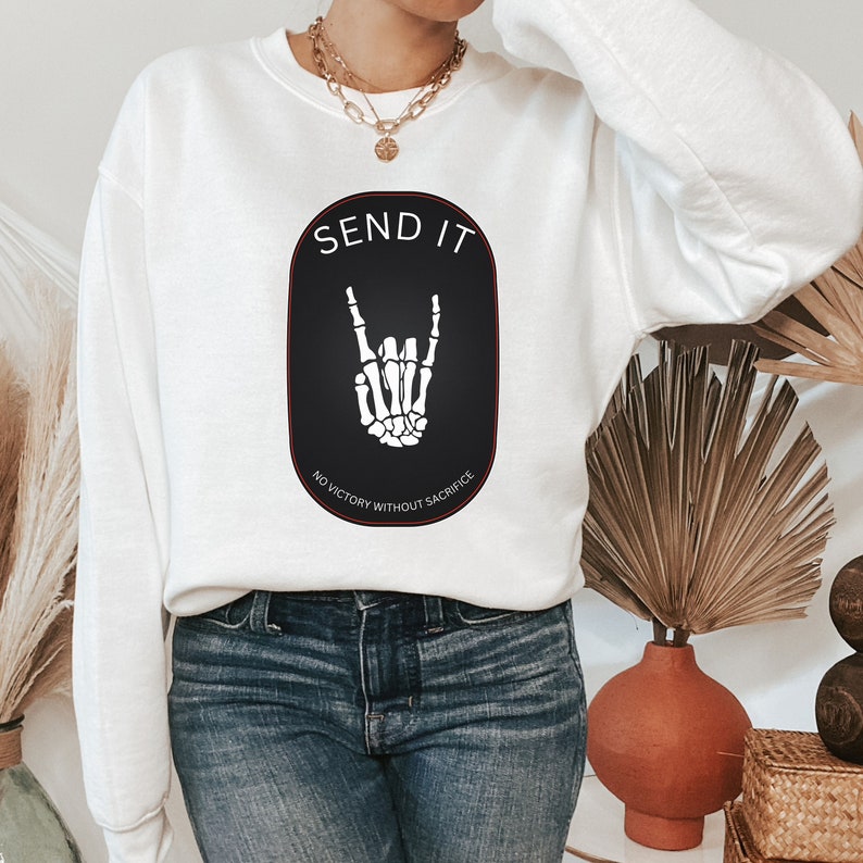 May include: White sweatshirt with a black oval graphic featuring a skeleton hand making the rock and roll sign. The text "SEND IT" is above the hand and "NO VICTORY WITHOUT SACRIFICE" is below the hand.
