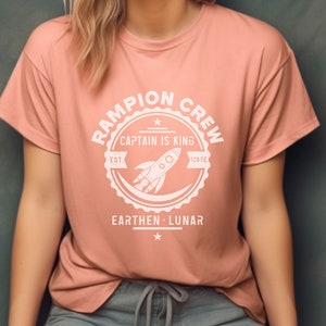May include: A pink t-shirt with a white graphic of a rocket ship and the text "Rampion Crew, Captain is King, Est. 126 T.E., Earthen - Lunar".