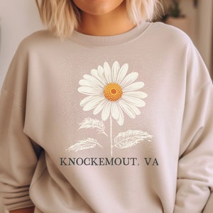 Puede incluir: Una sudadera beige claro con un gr&aacute;fico de margarita blanca y el texto "Knockemout, VA" impreso en la parte delantera.
