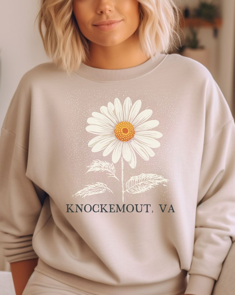 Puede incluir: Una sudadera beige claro con un gr&aacute;fico de margarita blanca y el texto "Knockemout, VA" impreso en la parte delantera.