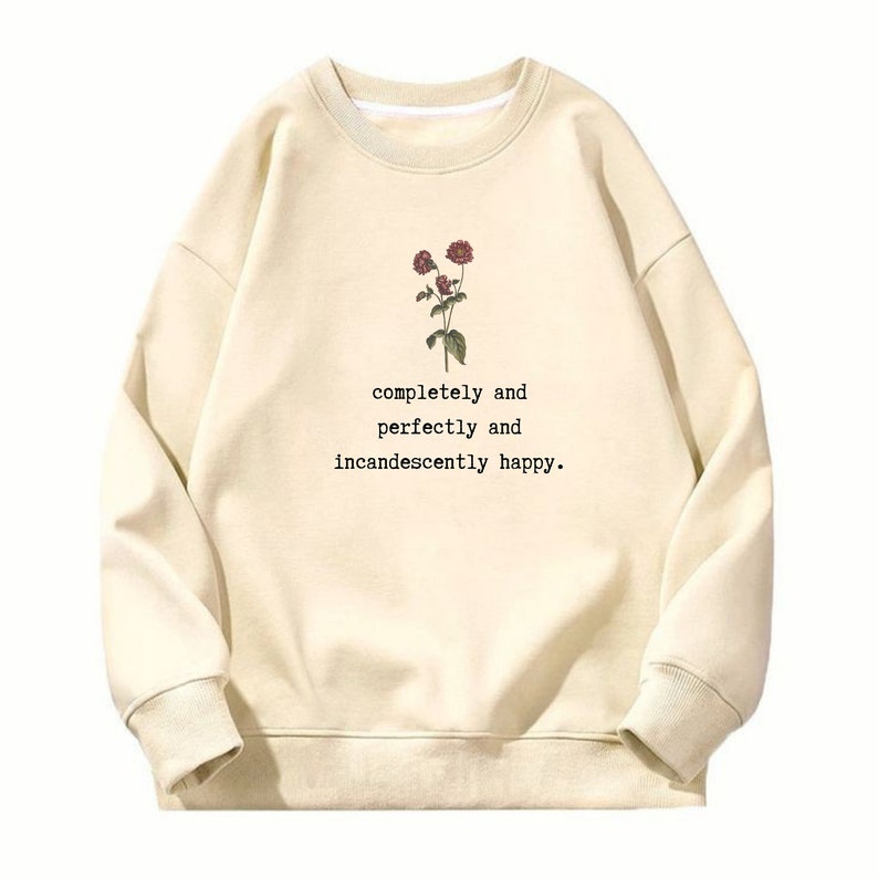 Puede incluir: Una sudadera de color crema con un gr&aacute;fico floral y el texto "completely and perfectly and incandescently happy."