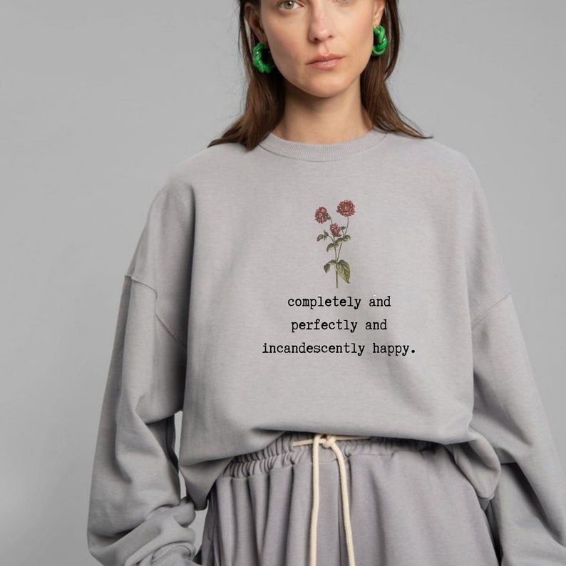 Puede incluir: Una sudadera gris claro con un gr&aacute;fico floral y el texto "completely and perfectly and incandescently happy."