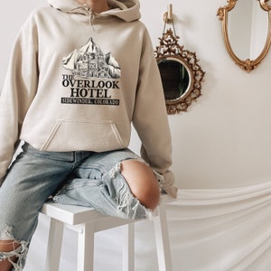 May include: A beige hoodie with a black graphic print of a mountain lodge with the text "The Overlook Hotel, Sidewinder, Colorado".