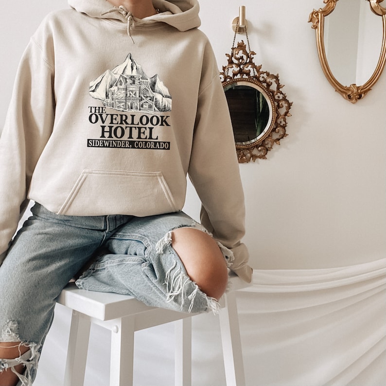 May include: A beige hoodie with a black graphic print of a mountain lodge with the text "The Overlook Hotel, Sidewinder, Colorado".