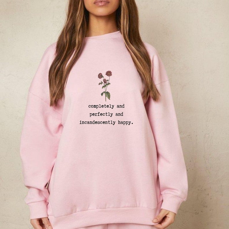 Puede incluir: Una sudadera rosa con un gr&aacute;fico floral y el texto "completely and perfectly and incandescently happy."