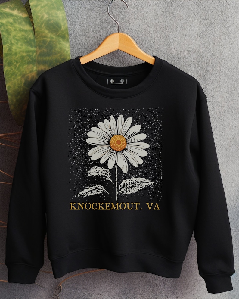 Puede incluir: Sudadera negra con un gr&aacute;fico de margarita blanca y el texto "Knockemout, VA" en amarillo.