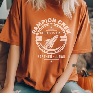 May include: A burnt orange t-shirt with a white graphic of a rocket ship inside a circle with the text "Rampion Crew, Captain is King, Est. 1261E, Earthen - Lunar".