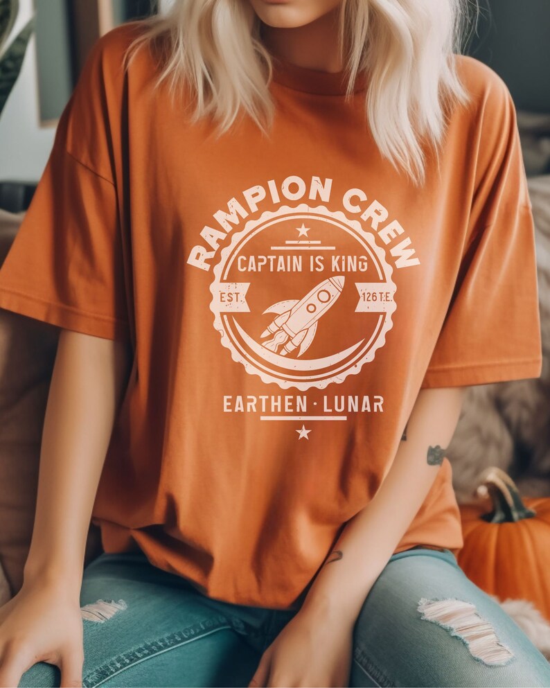 May include: A burnt orange t-shirt with a white graphic of a rocket ship inside a circle with the text "Rampion Crew, Captain is King, Est. 1261E, Earthen - Lunar".