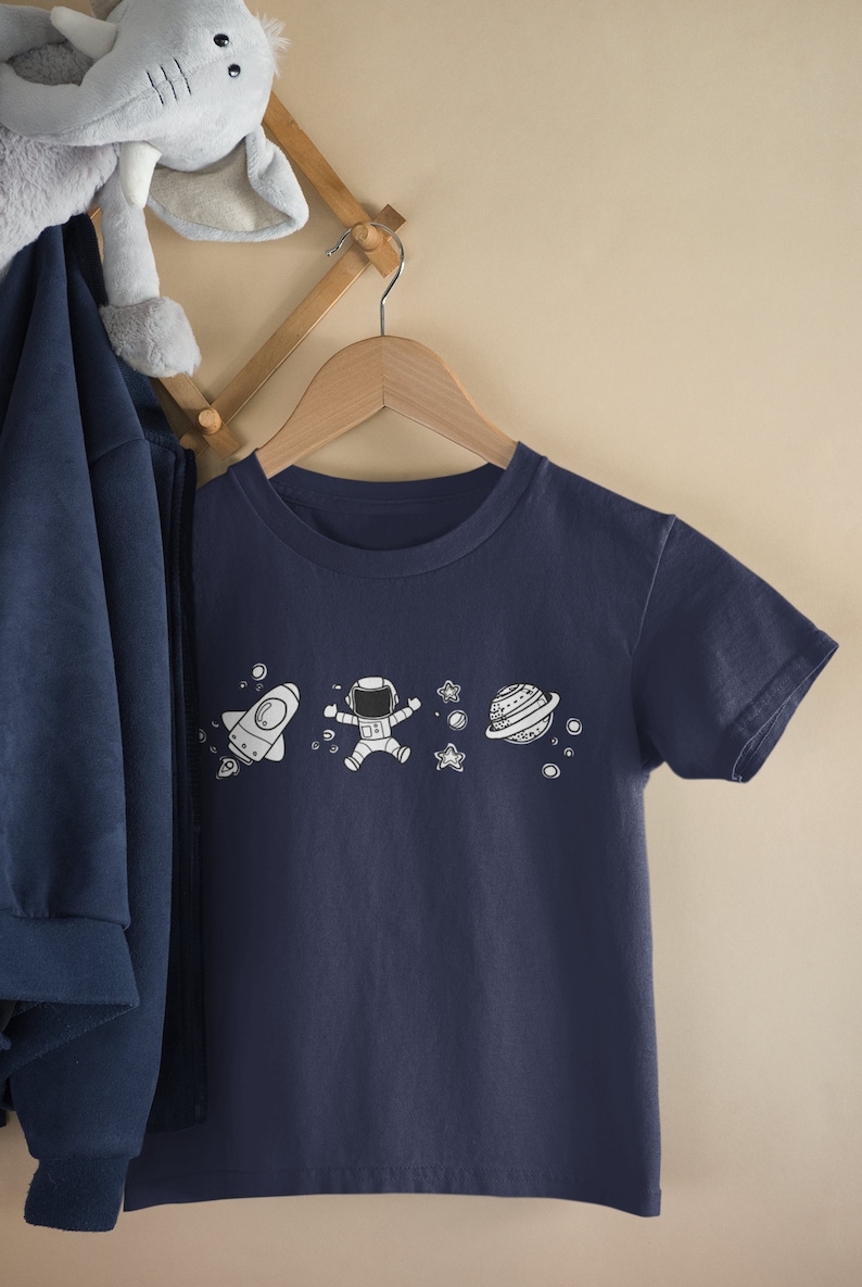 May include: A navy blue t-shirt with a white astronaut, rocket, and planets graphic. The shirt is hanging on a wooden hanger.