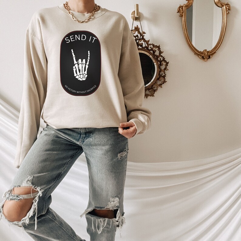 May include: A beige sweatshirt with a black graphic of a skeleton hand making a rock and roll sign. The graphic is inside a black oval with the text "SEND IT" above the graphic and "NO VICTORY WITHOUT SACRIFICE" below the graphic.