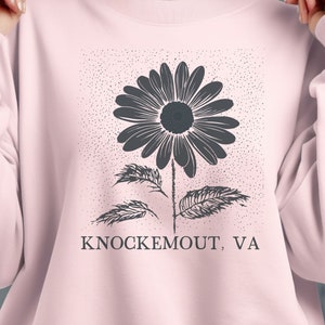 Puede incluir: Una sudadera rosa con un gr&aacute;fico en blanco y negro de una flor y el texto "Knockemout, VA".