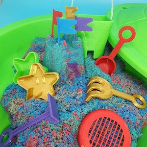May include: A green plastic sandbox filled with colorful kinetic sand. The sandbox includes a green bucket, a red shovel, a gold rake, a purple tool, a red sieve, a gold star-shaped mold, and four colorful flags.