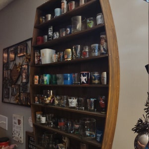 Może przedstawiać: Drewniana półka w kształcie beczki, wypełniona różnymi kieliszkami do wódki. Półka jest w kolorze ciemnobrązowym i zamontowana na szarej ścianie. Kieliszki mają różne rozmiary i wzory, niektóre z obrazami i tekstem.