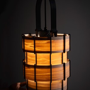 Pu&ograve; includere: Un lampadario cilindrico con un interno caldo e luminoso. La luce &egrave; realizzata in legno a strati e supporti in metallo nero. Il lampadario &egrave; sospeso a un cavo nero e ha una maniglia in metallo nero nella parte superiore.