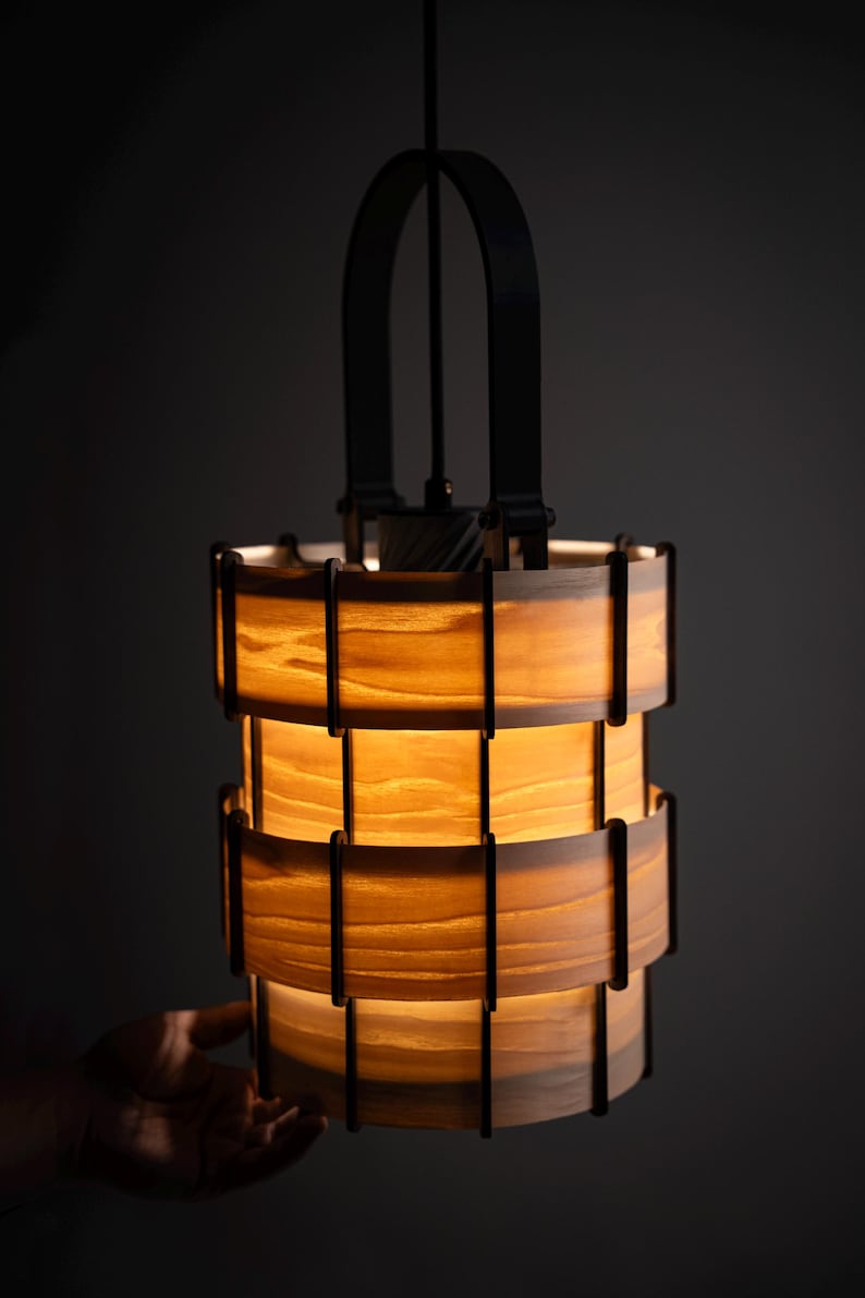 Pu&ograve; includere: Un lampadario cilindrico con un interno caldo e luminoso. La luce &egrave; realizzata in legno a strati e supporti in metallo nero. Il lampadario &egrave; sospeso a un cavo nero e ha una maniglia in metallo nero nella parte superiore.