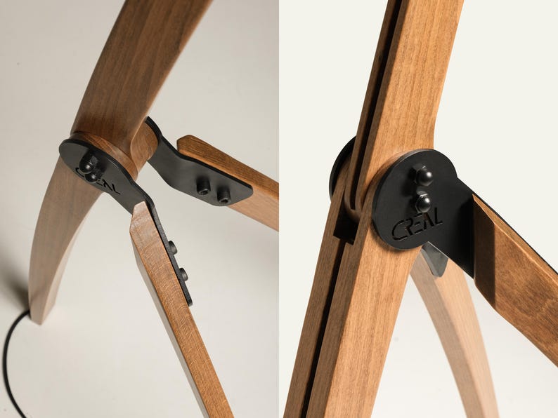 Pu&ograve; includere: Primo piano di una lampada a treppiede in legno con cerniere e dettagli in metallo nero. Le gambe in legno della lampada sono marrone chiaro e curve, collegate da piastre metalliche nere. La parola "CREAL" &egrave; visibile.