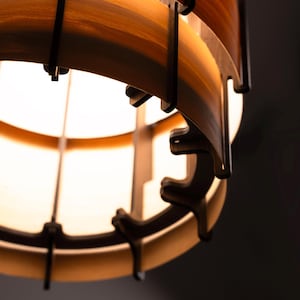 Pu&ograve; includere: Primo piano di un lampadario in legno. Il lampadario presenta doghe di legno a strati in tonalit&agrave; marroni e beige, tenute insieme da supporti marrone scuro. La sorgente luminosa &egrave; visibile all'interno, creando un bagliore caldo.