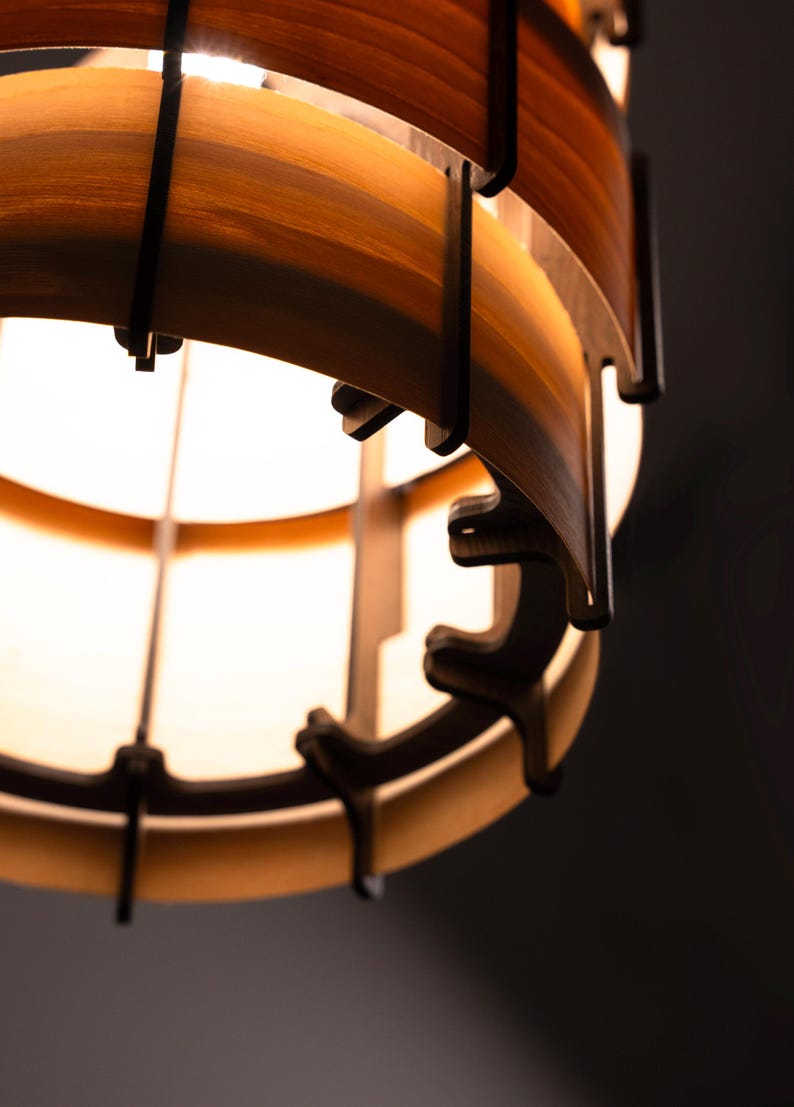 Pu&ograve; includere: Primo piano di un lampadario in legno. Il lampadario presenta doghe di legno a strati in tonalit&agrave; marroni e beige, tenute insieme da supporti marrone scuro. La sorgente luminosa &egrave; visibile all'interno, creando un bagliore caldo.