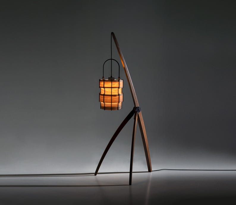 Pu&ograve; includere: Una lampada da terra moderna con una base a treppiede in legno e un braccio di supporto curvo. La lampada presenta un paralume cilindrico realizzato con doghe di legno impilate, con una luce calda e brillante. L'altezza della lampada &egrave; di circa 152 cm.