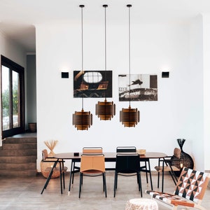May include: Three brown wooden pendant lights hang over a dining table with six chairs. The lights are made of thin wooden slats and have a modern design. The table is made of wood and has a natural finish. The chairs are upholstered in brown leather and have black metal frames.