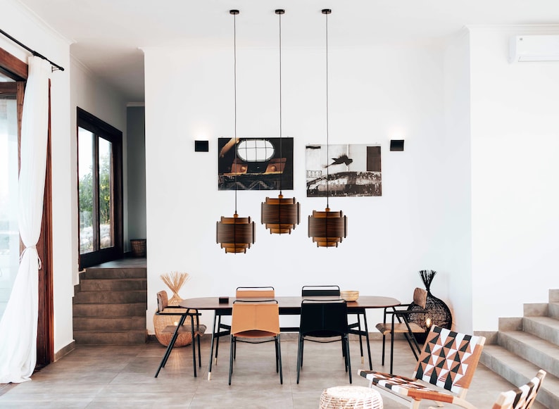 May include: Three brown wooden pendant lights hang over a dining table with six chairs. The lights are made of thin wooden slats and have a modern design. The table is made of wood and has a natural finish. The chairs are upholstered in brown leather and have black metal frames.