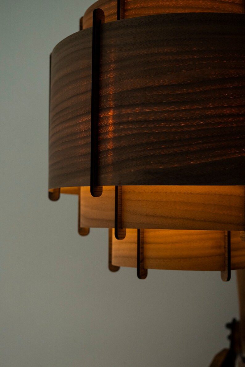 May include: A wooden pendant light fixture with multiple layers of wood, each with a different grain pattern. The top layer is dark brown, and the bottom layer is a lighter brown. The light is turned on, and the light shines through the wood.