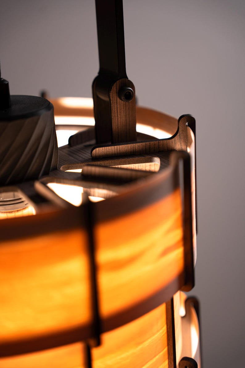 Pu&ograve; includere: Primo piano di una lampada in legno con una calda luce arancione. La lampada presenta un design a strati con dettagli in legno marrone scuro e un braccio orientabile. Il design &egrave; moderno e minimalista, adatto all'arredamento.