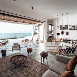 May include: A living room with large windows overlooking the ocean. The room is decorated in a modern style with a white sofa, wicker chairs, and a woven rug. There are several hanging lights and a large coffee table with a woven top.