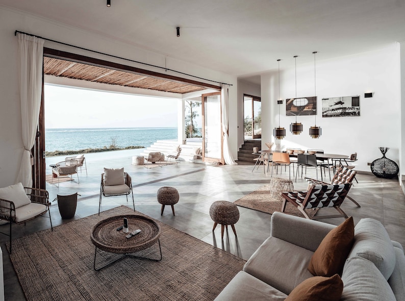 May include: A living room with large windows overlooking the ocean. The room is decorated in a modern style with a white sofa, wicker chairs, and a woven rug. There are several hanging lights and a large coffee table with a woven top.