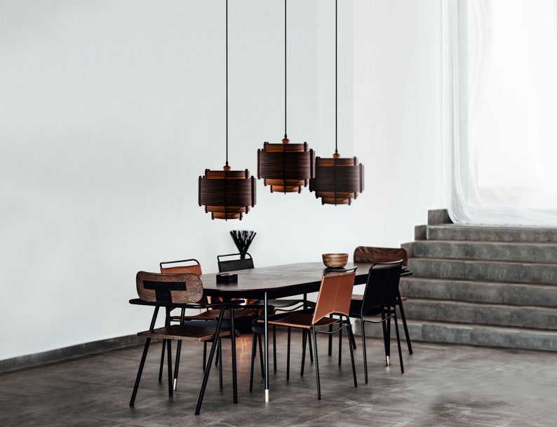 May include: Three brown wooden pendant lights hang over a dining table with six chairs. The table is made of dark wood and the chairs have black metal frames and brown leather seats. The room has a white wall and a concrete staircase in the background.
