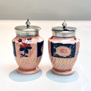 May include: Two antique pink ceramic jars with silver lids. Each jar features a dark blue rectangular panel with floral designs and gold accents. The jars have a bulbous shape and are likely for spices or condiments.