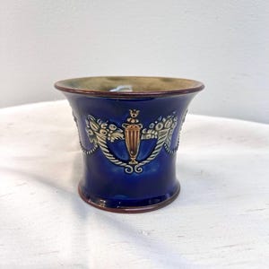 May include: A dark blue ceramic planter with a decorative gold and white floral design. The planter has a flared rim and a brown rim and base. The design includes a central urn-like shape.