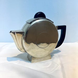May include: A circular silver-toned metal teapot featuring a flat, disc-shaped body with a central circular indentation. It includes a short, angular spout, a black handle, and a black circular knob on the lid, all resting on a small, square base.