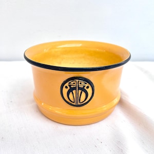 May include: A small, round, yellow ceramic bowl with a black rim and a black circular emblem on the side. The emblem features an abstract design. The bowl is sitting on a white surface.