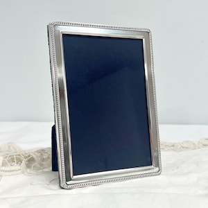 May include: A rectangular picture frame with a polished silver-tone metallic finish and a beaded border detail. The frame features a dark navy blue insert and a black velvet backing, designed to stand upright on a flat surface for display.
