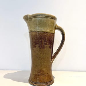 Op de afbeelding: Een hoge, handgemaakte keramische kan met een brede basis en een gebogen handvat. De kan heeft een kleurovergang, van licht olijfgroen aan de bovenkant naar een diep bruin aan de basis. De kan is ongeveer 30 cm hoog.